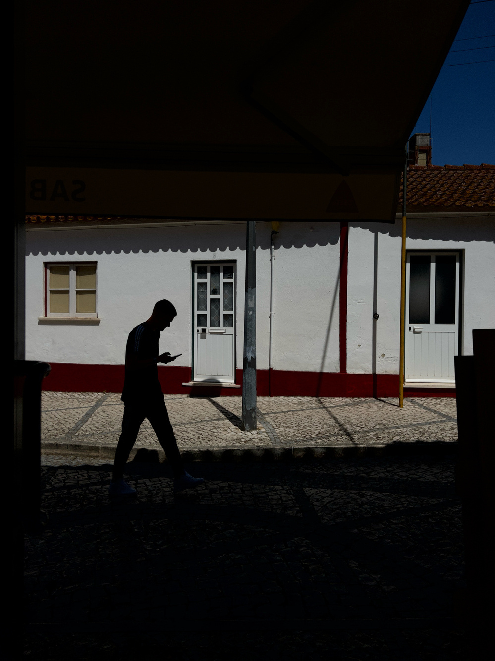 Silhouette of a person walking along a cobblestone street, holding a smartphone. The street is lined with white buildings with red and yellow trim in the background, and the scene is partially shaded.