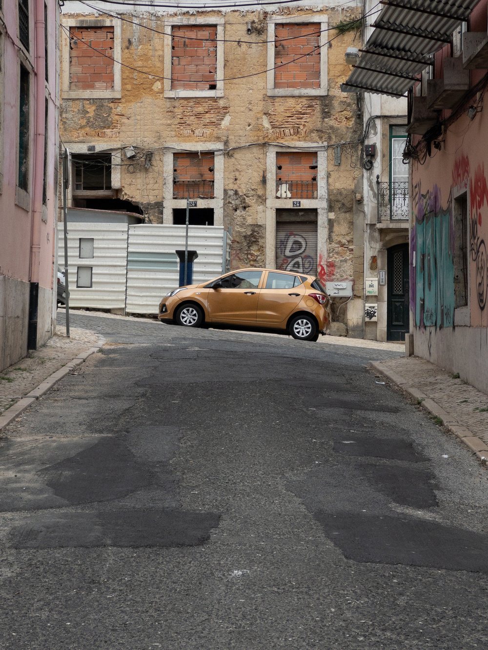 A narrow, slightly inclined street with a worn and patched asphalt surface, leading to an old, abandoned building with boarded-up windows. A bronze-colored car is parked horizontally across the street near the building. Surrounding walls feature graffiti and some exposed brick.