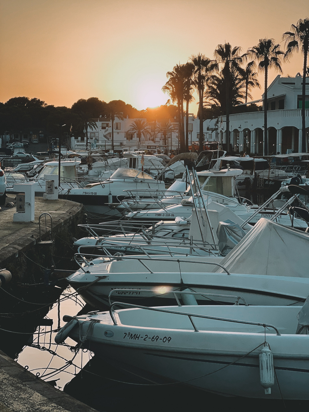 A serene marina at sunset with several moored boats and yachts. Palm trees and white buildings are visible in the background, under a sky transitioning from golden to dark.