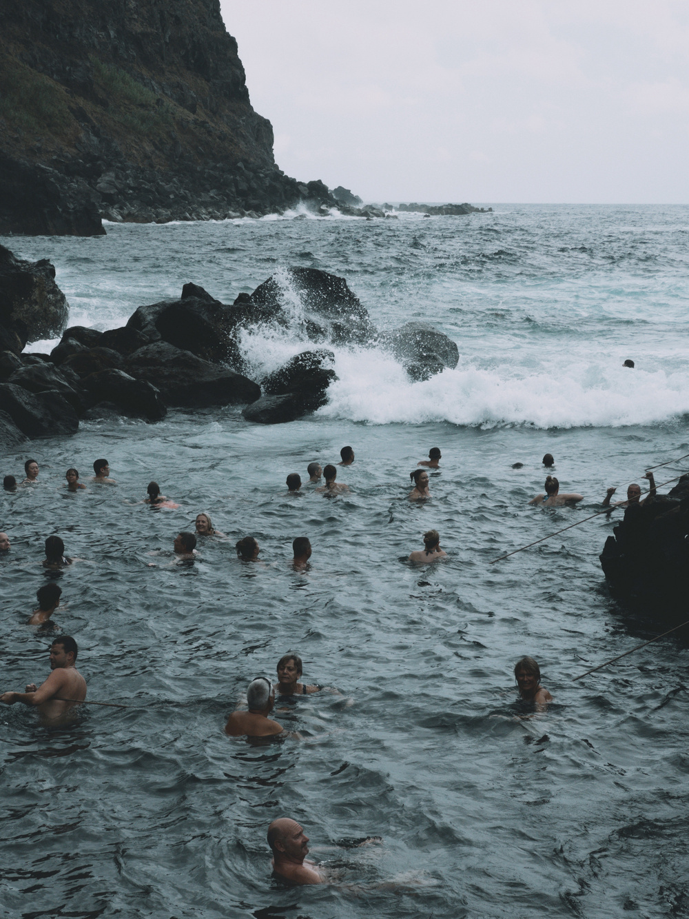 A rocky coastline with waves crashing against the rocks. Many people are swimming and enjoying the water near the shore, holding onto ropes anchored on the rocks for support. Lush, green foliage is visible on the cliffside in the background. Overcast sky.