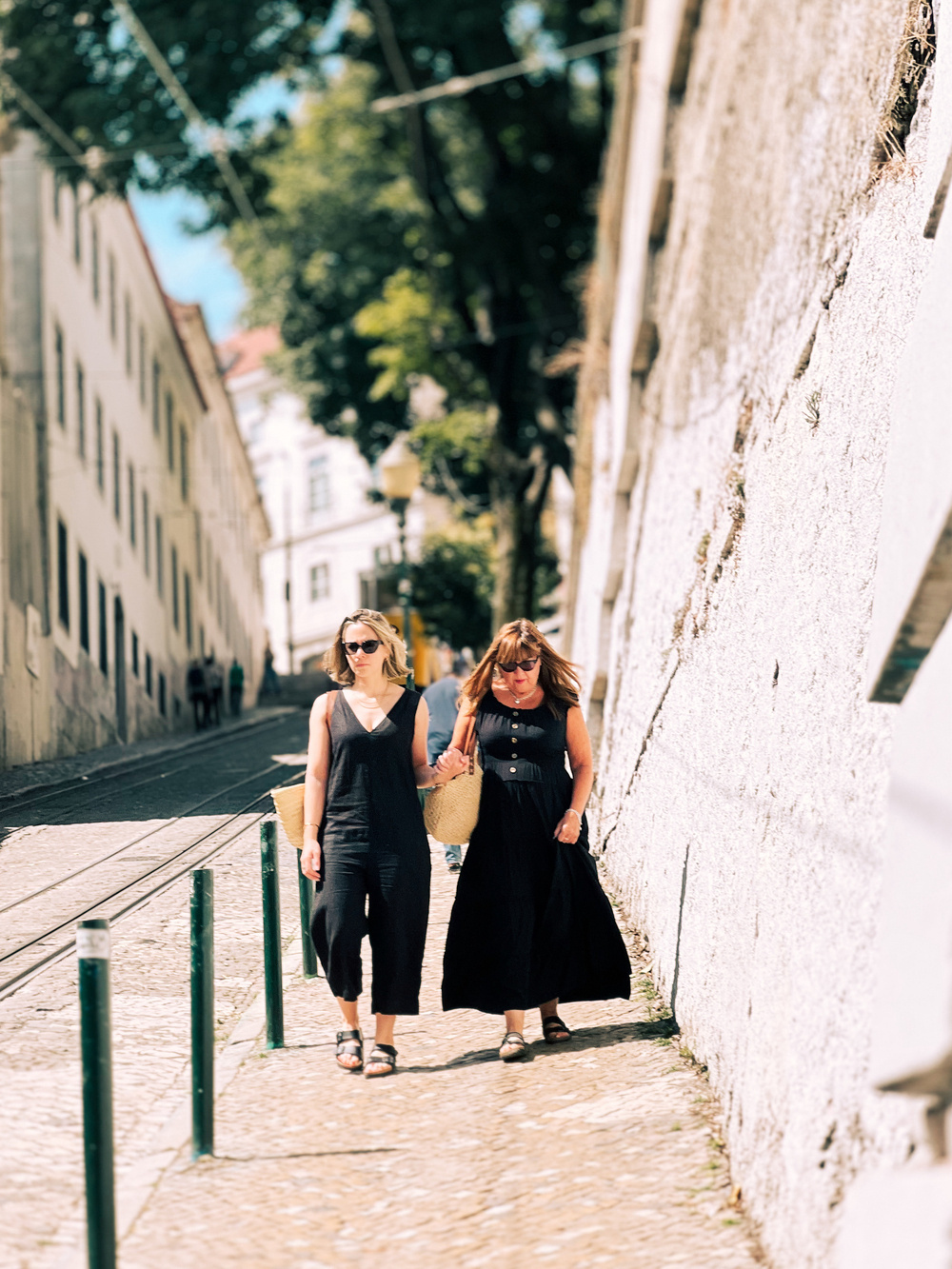 Two women in black dresses walk arm-in-arm along a cobblestone street lined with buildings. They are carrying woven bags and wearing sunglasses. The street is bordered by green poles and has tram tracks running through it.