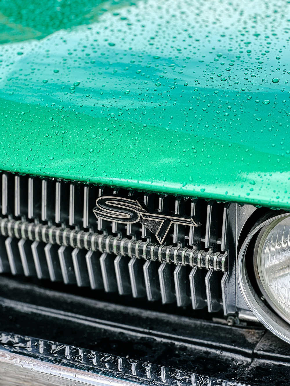 Close-up of a green car&rsquo;s front end with water droplets, featuring the grille and an &ldquo;ST&rdquo; badge next to a headlight.