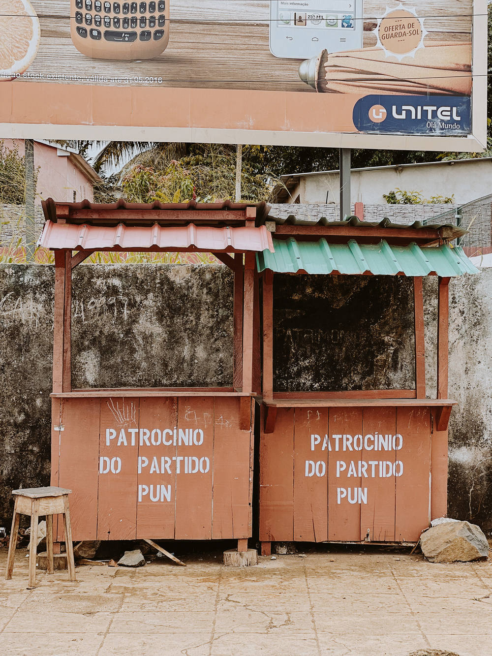 The image shows two small wooden market stalls with red and green roofs. Both stalls have the text “PATROCINIO DO PARTIDO PUN” painted on them. A wooden stool is placed in front of one stall.