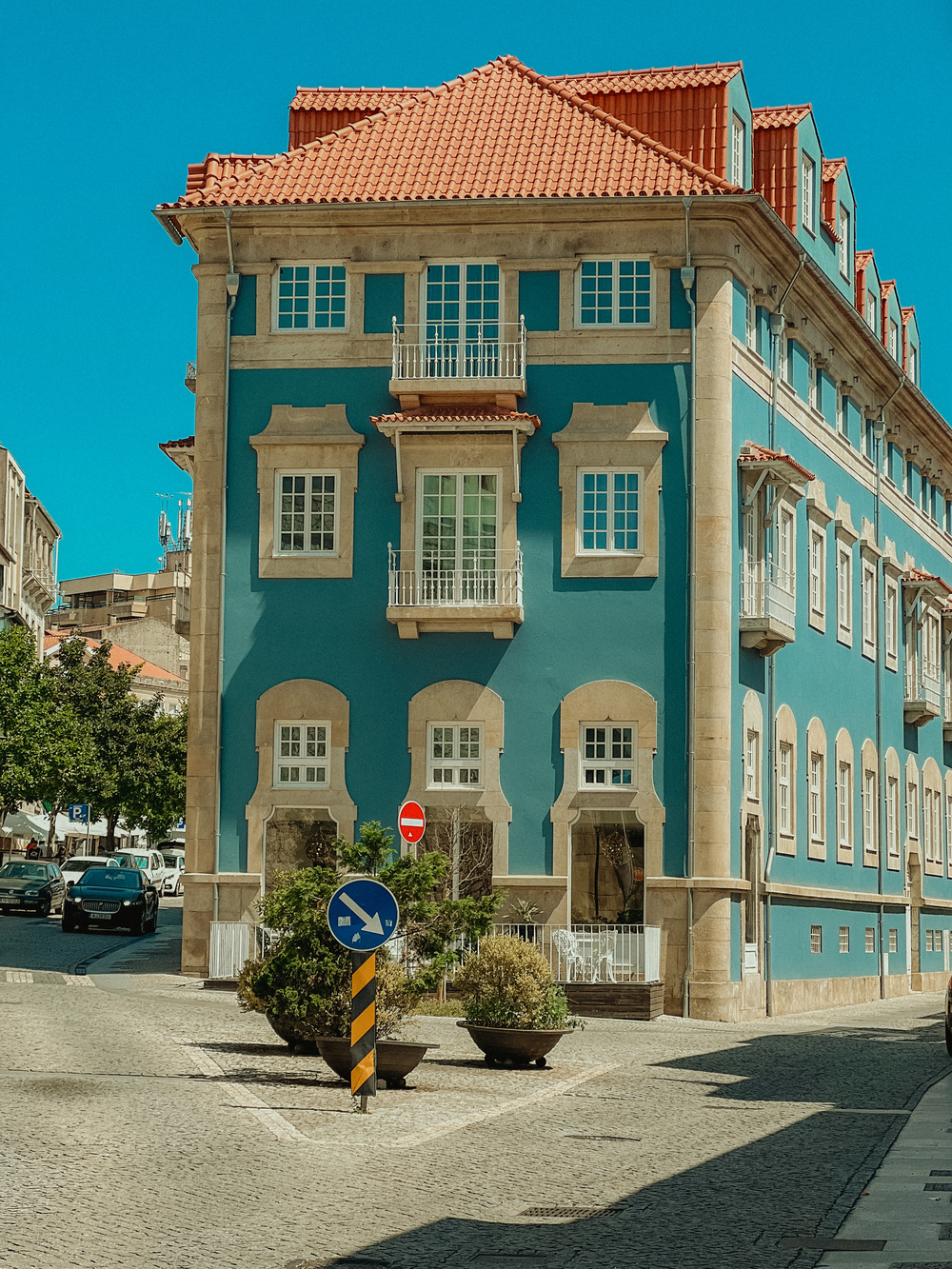 A vibrant blue building with beige stone accents and a red roof stands prominently on a cobblestone street. The building features multiple windows, small balconies, and a ground floor that appears to house a café or restaurant. There are several potted plants.