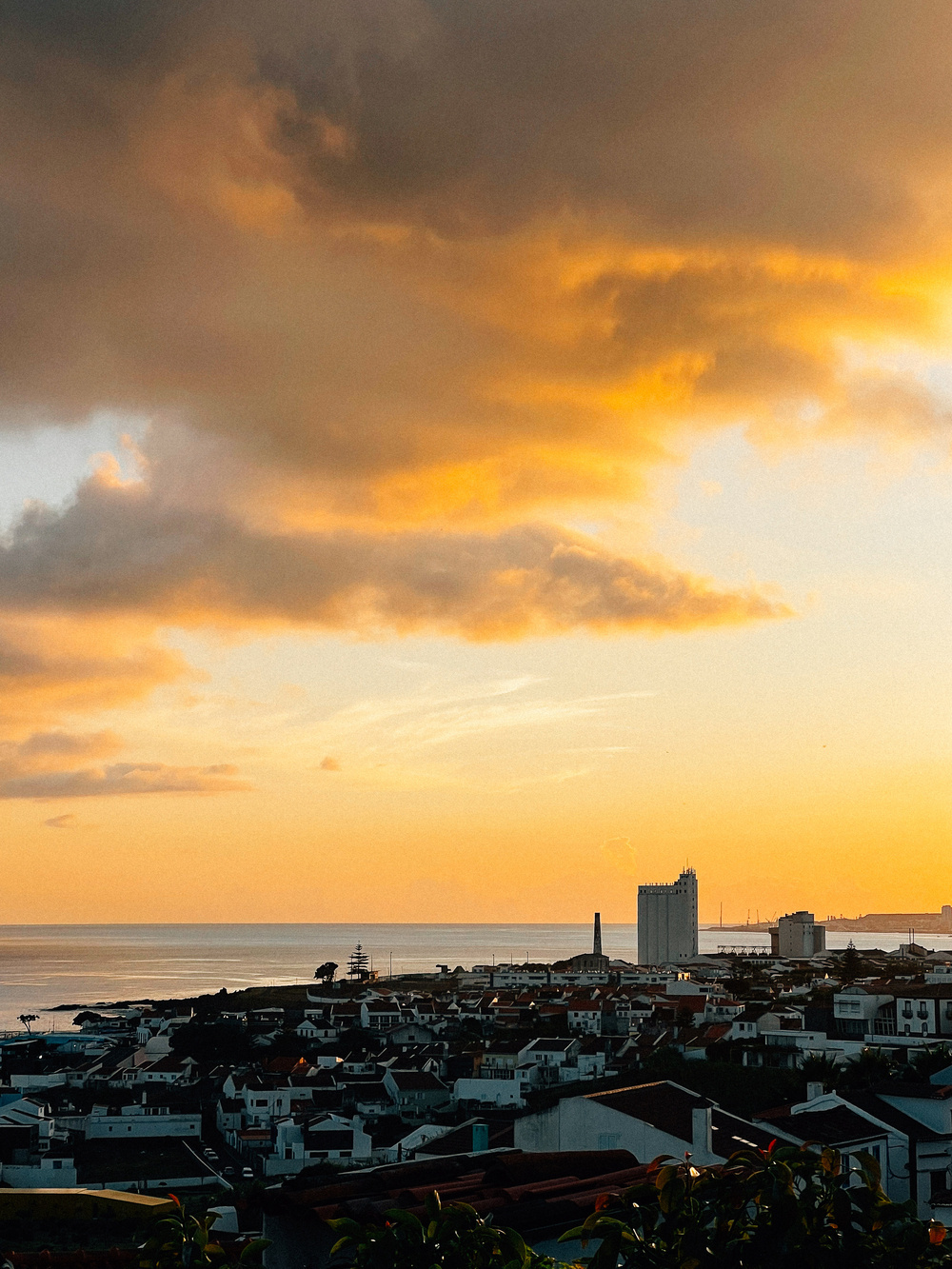 A cityscape at sunset overlooking a coastal area with buildings silhouetted against the orange and golden sky. A tall building and a chimney-like structure stand out in the distance. The tranquil sea is visible in the background.