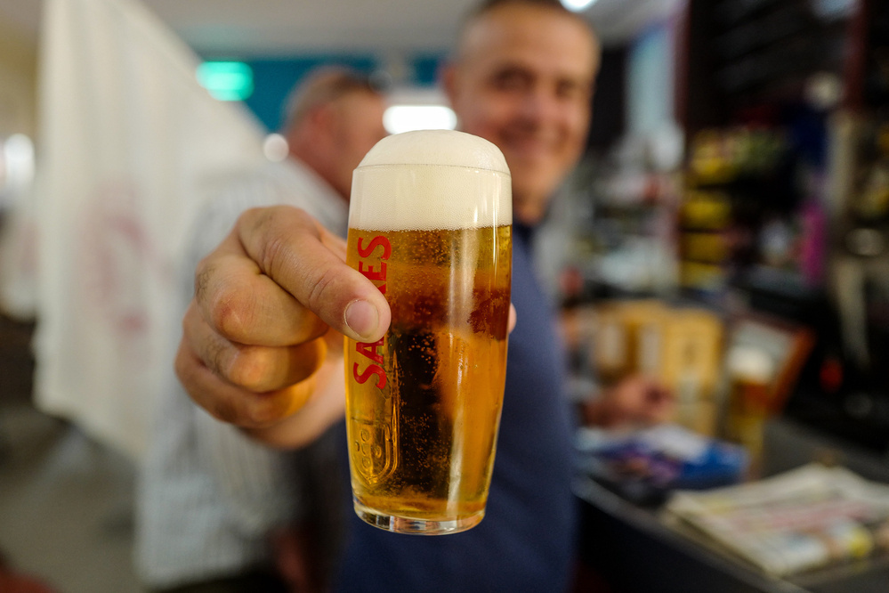 A person holding a glass of beer with foam on top, labeled “SAGRES” in red letters. The background is out of focus, showing another individual and miscellaneous items in what appears to be a casual indoor setting.