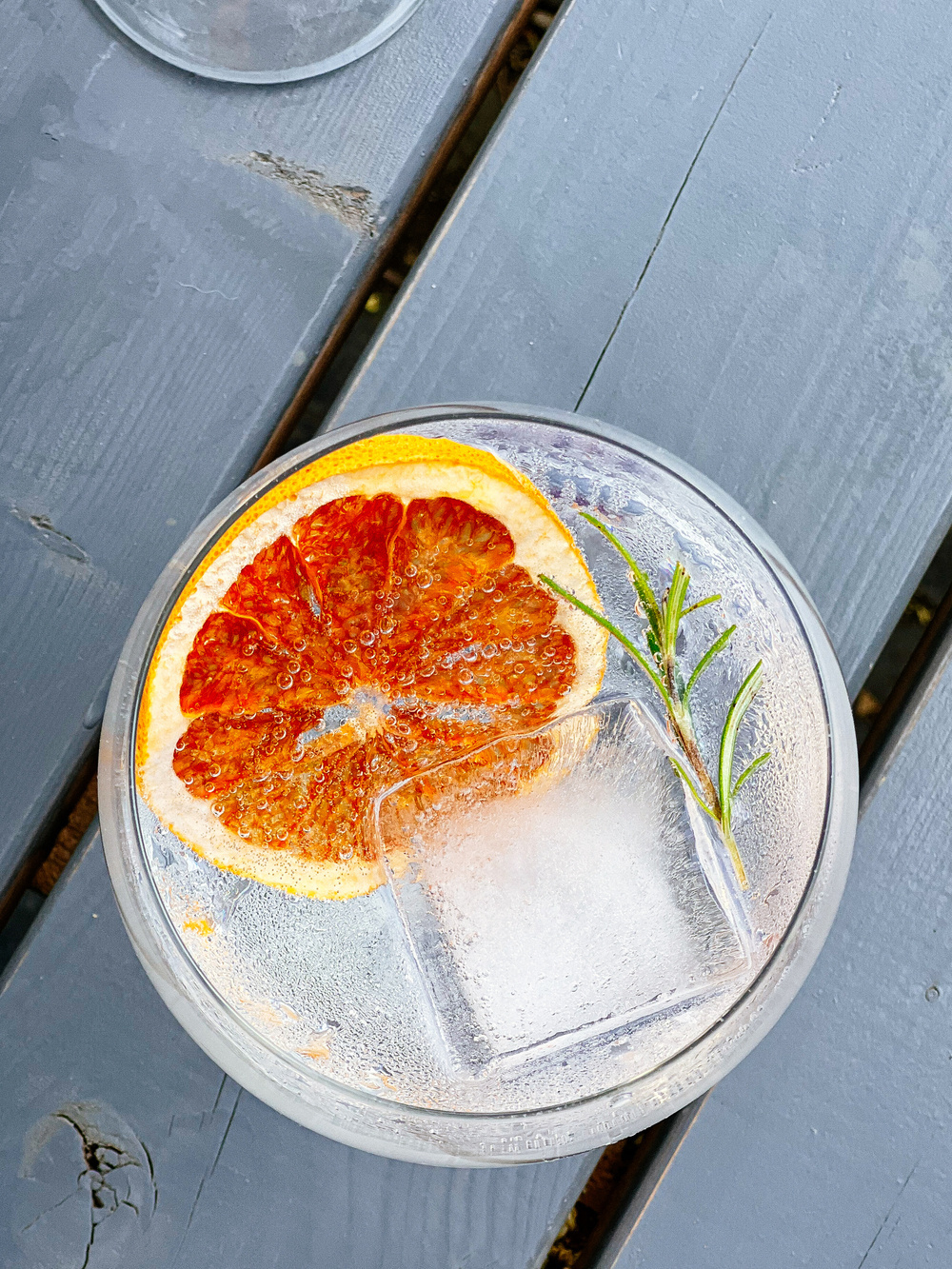 A top view of a refreshing drink in a glass, garnished with a slice of blood orange and a sprig of rosemary. The drink contains ice cubes and is placed on a gray wooden surface.