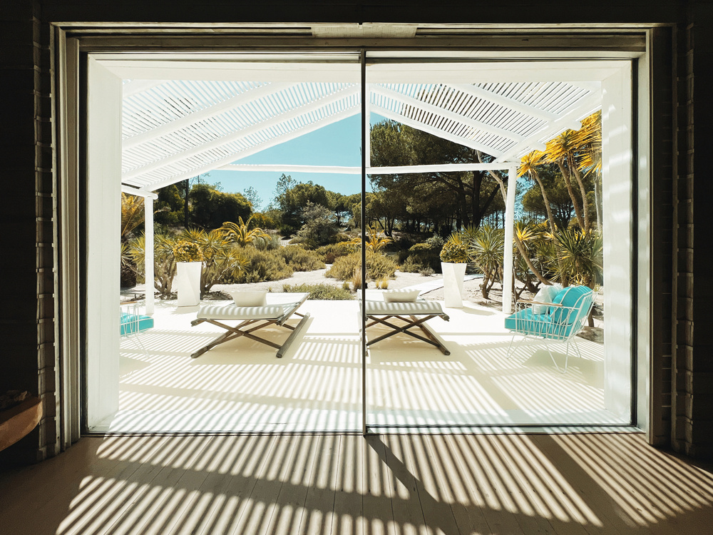 Image of a patio viewed through a large glass door, featuring an outdoor seating area with a lounger and blue chair. The patio is shaded by a pergola with slatted white panels casting striped shadows. In the background, there are various plants.