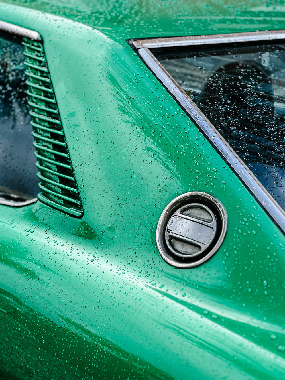 Close-up of a teal car with raindrops on the surface, featuring a visible side vent and a car manufacturer&rsquo;s logo badge.