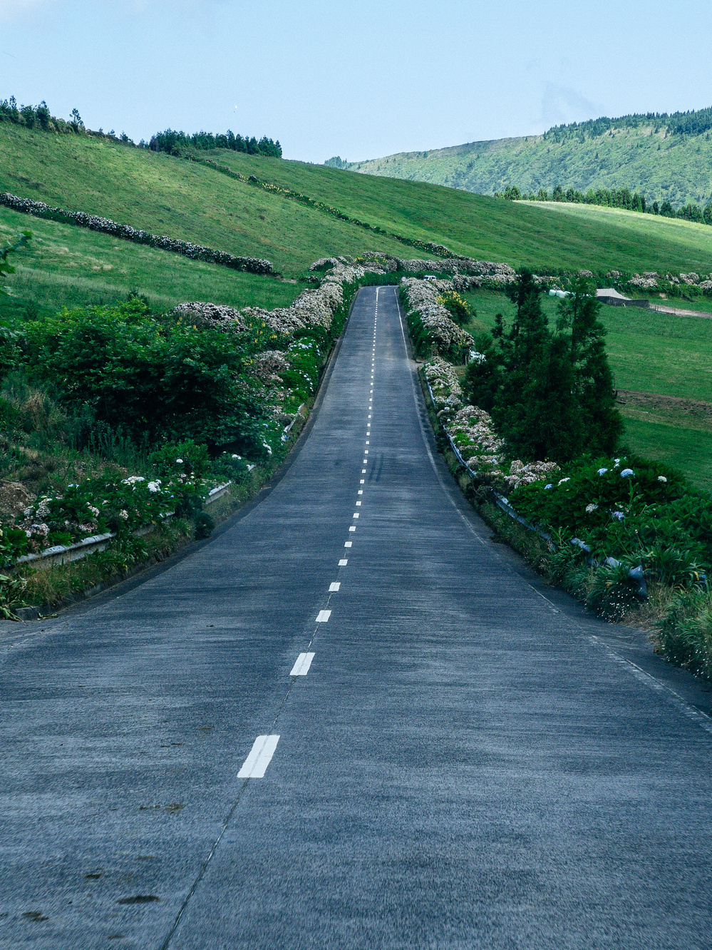 A long, straight road stretches into the distance through green, rolling hills. Stone walls and dense vegetation line the sides of the road. The sky is blue with a few scattered clouds.