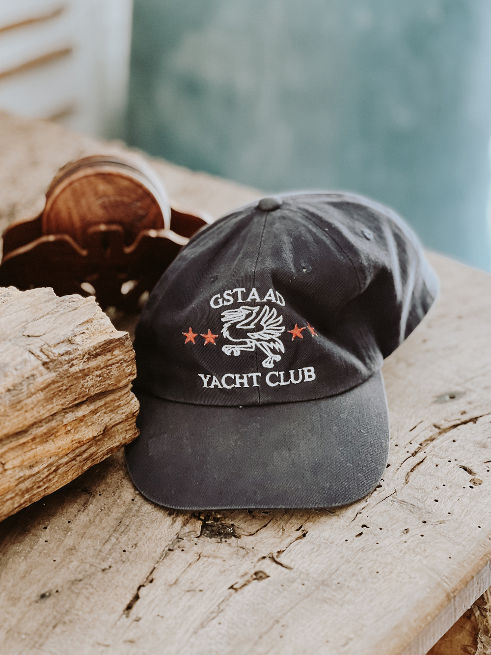 A black baseball cap is placed on a rustic wooden surface. The cap features an embroidered design with the words &ldquo;GSTAAD YACHT CLUB&rdquo; along with an eagle and three red stars. Behind the cap, there is a wooden object. 