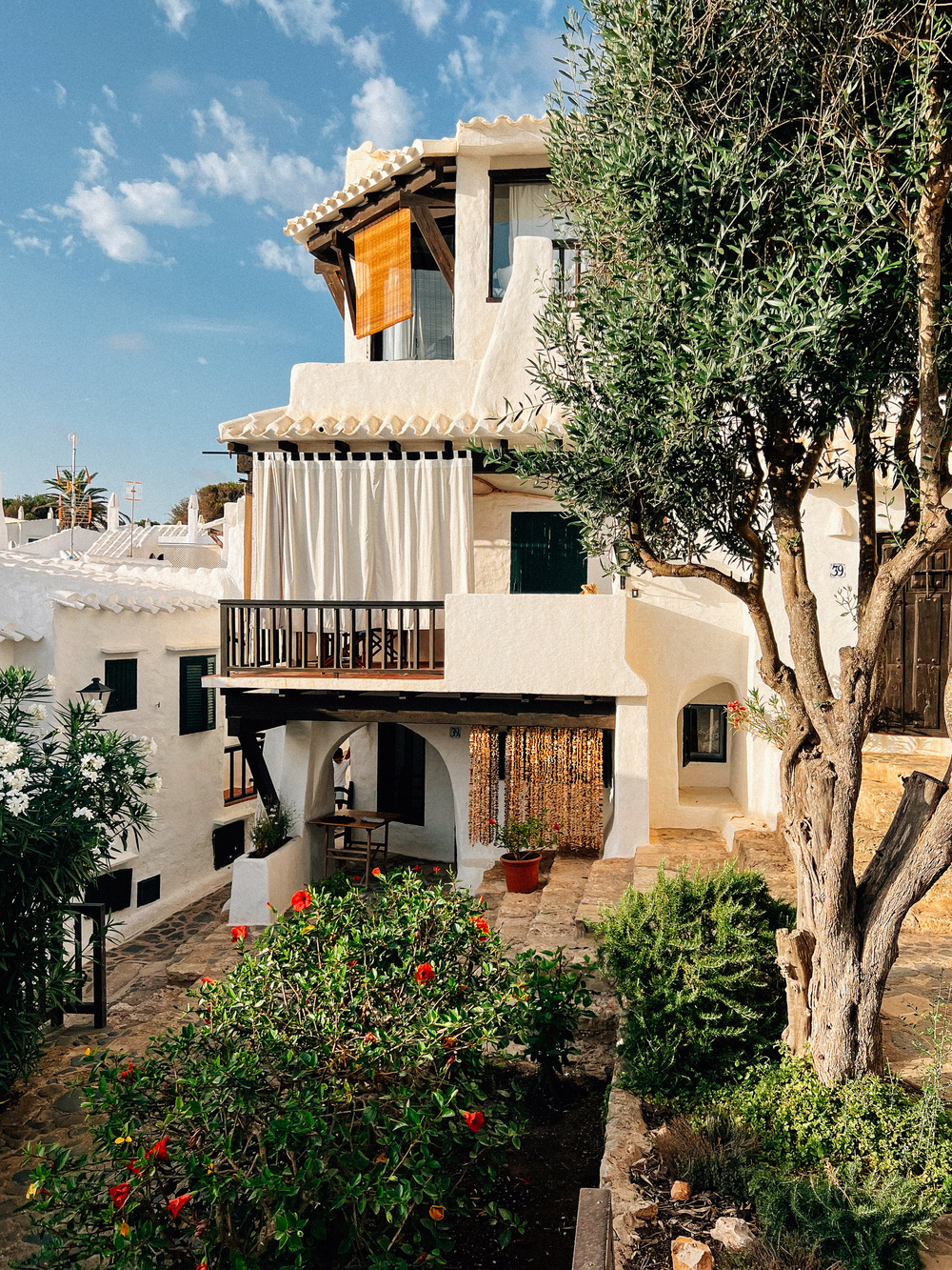 A charming Mediterranean-style house with white stucco walls and terracotta roof tiles. The house features a wooden balcony with draped curtains and beaded doorway curtains. A lush garden with flowering plants and trees surrounds the stone pathway leading to the entrance.