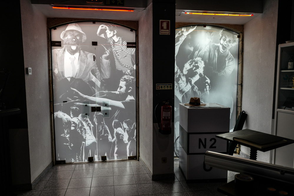 The image shows a hallway with two large doors covered in black and white mural art depicting various people in dramatic gestures. A fire extinguisher hangs on a column in the center, with a display counter on the right side that has a hat placed on top of it. 