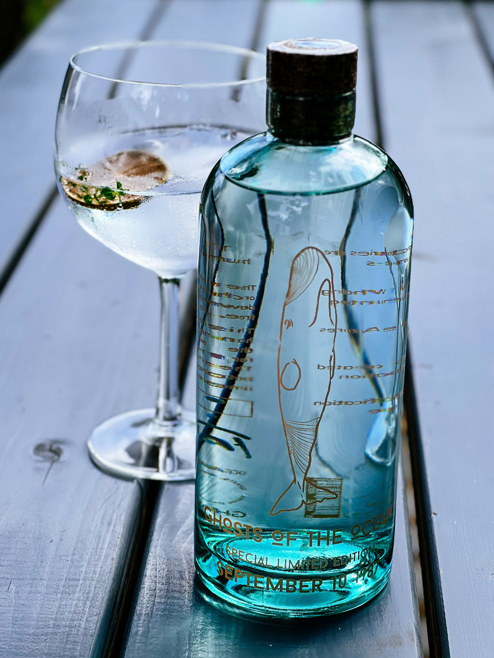 A clear, blue-tinted glass bottle labeled &ldquo;GHOSTS OF THE OCEAN (SPECIAL LIMITED EDITION SEPTEMBER 10)&rdquo; is placed on a wooden table beside a large, round glass containing a transparent beverage with ice and a garnish.