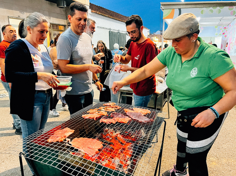 People at an outdoor event with a woman grilling meat on a barbecue, and others waiting to be served or already with food on their plates.
