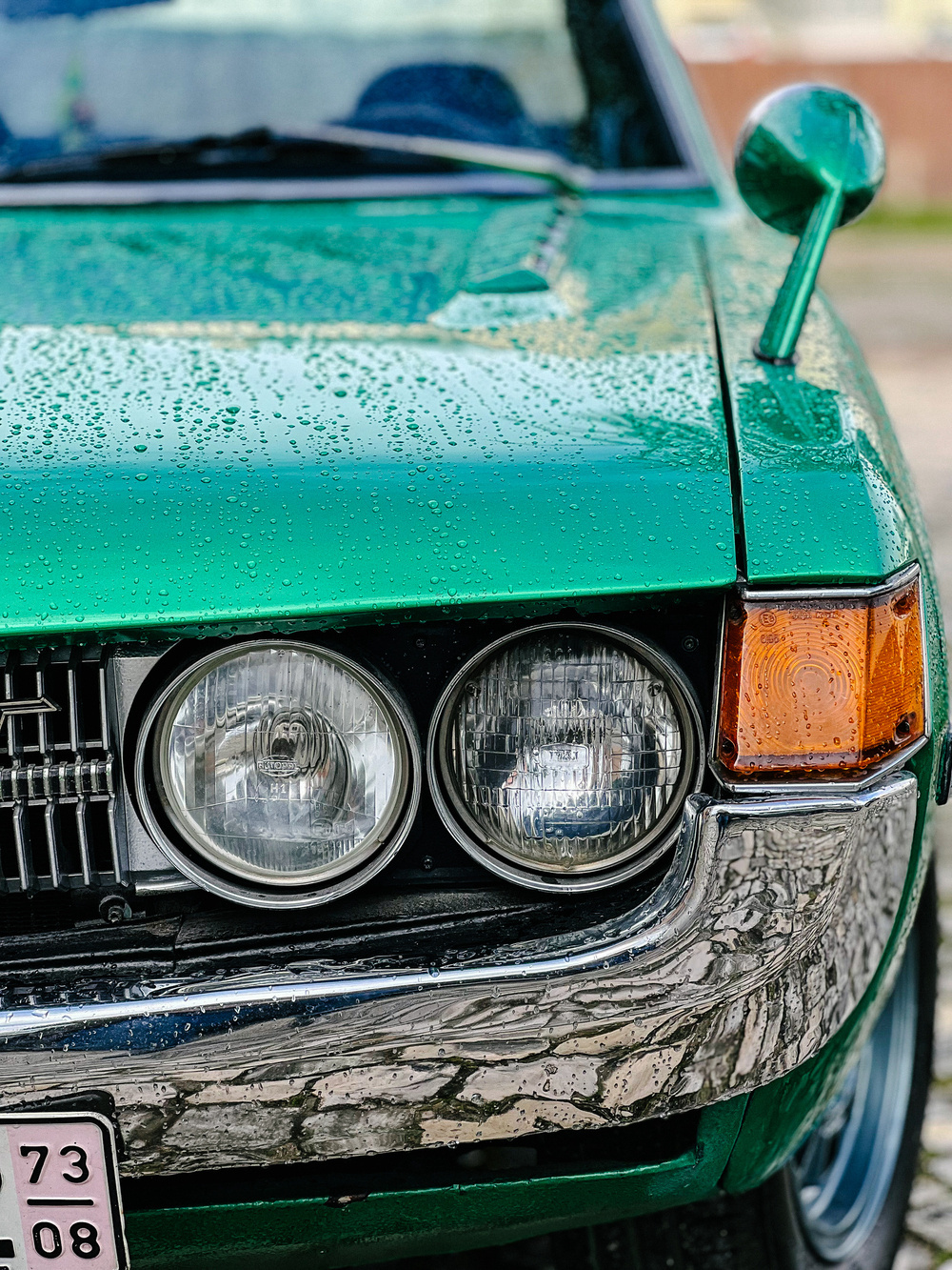 A close-up of a green vintage car&rsquo;s front section with a focus on the grille, headlamps, and turn signal, covered in raindrops.