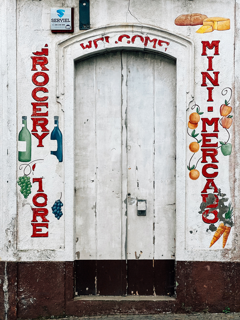 This image shows the entrance to a grocery store or mini-market. The doorway is framed by murals of fruits, vegetables, bottles, bread, and cheese, with the words &ldquo;GROCERY STORE&rdquo; on the left and &ldquo;MINI MERCADO”.