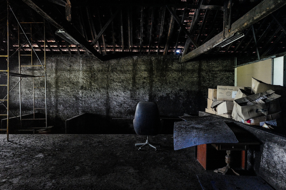 A dimly lit, abandoned room with a dark, rocky wall and exposed wooden roof beams. An empty black office chair sits in the center. There is a yellow metal ladder on the left and a pile of broken-down cardboard boxes and debris on the