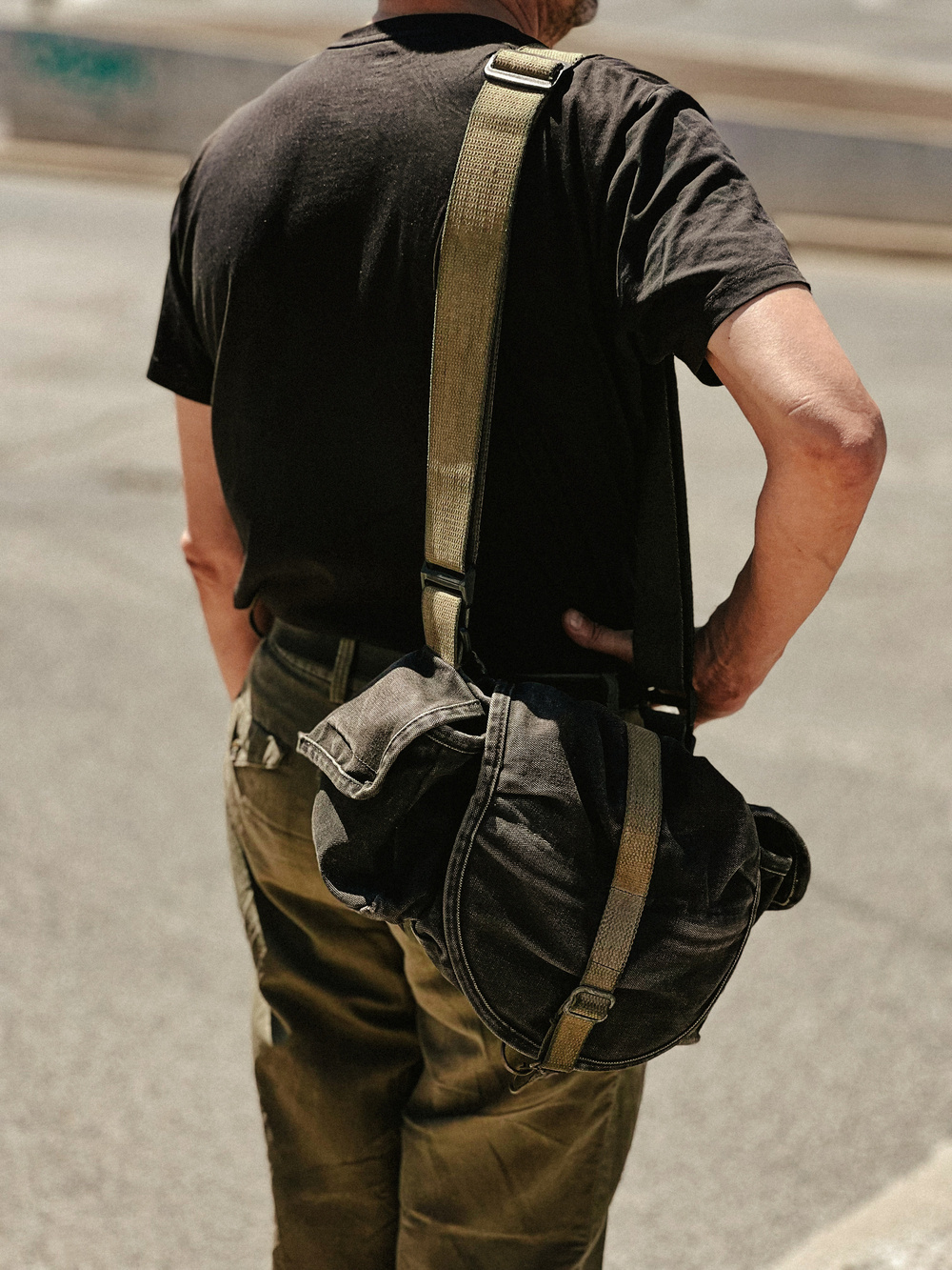 A person wearing a black T-shirt and olive green pants is seen from behind, carrying a black shoulder bag with a tan strap over their left shoulder. The background is an urban street setting.