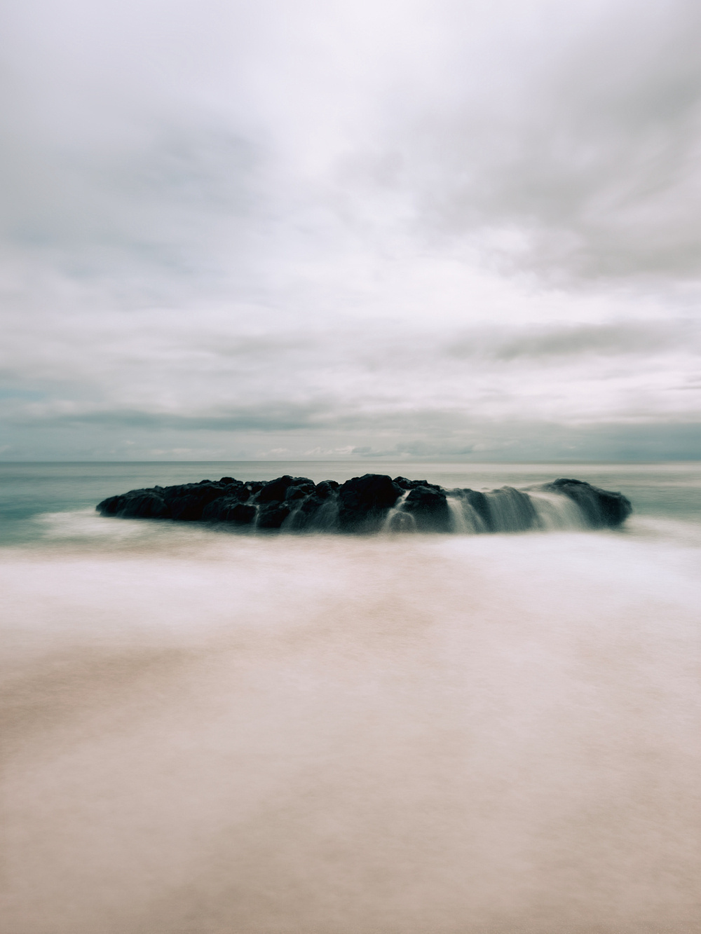 A long exposure photograph of waves crashing over an isolated rock formation on a beach, with a cloudy sky in the background. The image has a serene and ethereal quality due to the soft, blurred water and smooth sand.