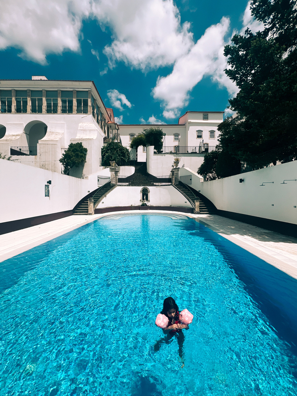 A child wearing pink inflatable armbands swims in a clear blue pool. The pool is situated in front of a large white building with arches and multiple windows. The sky above is partly cloudy.