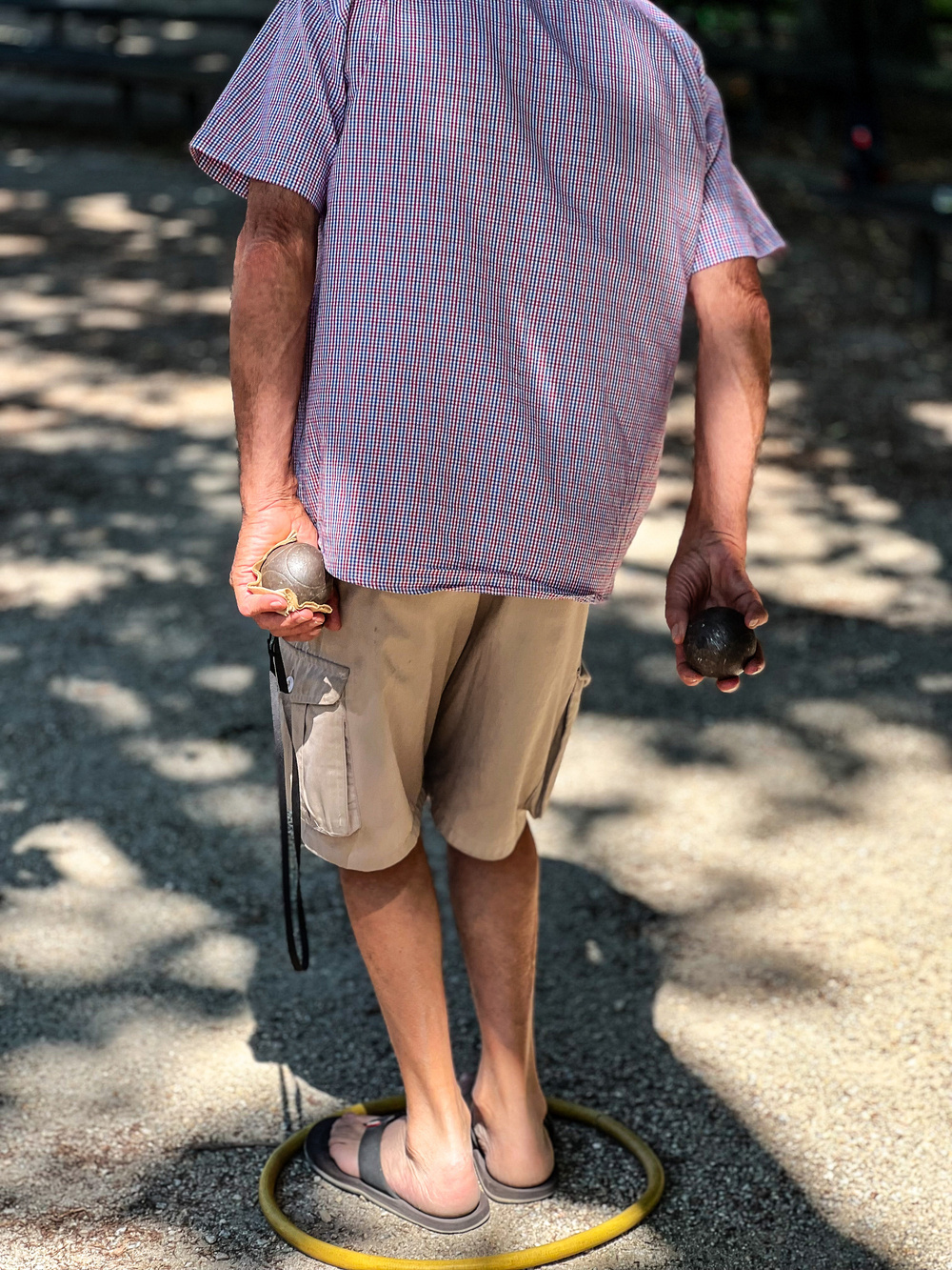 A person, seen from behind, stands inside a yellow circle on a gravel surface. They are holding metal boules, and they are wearing a short-sleeved, checkered shirt, beige shorts, and black sandals.