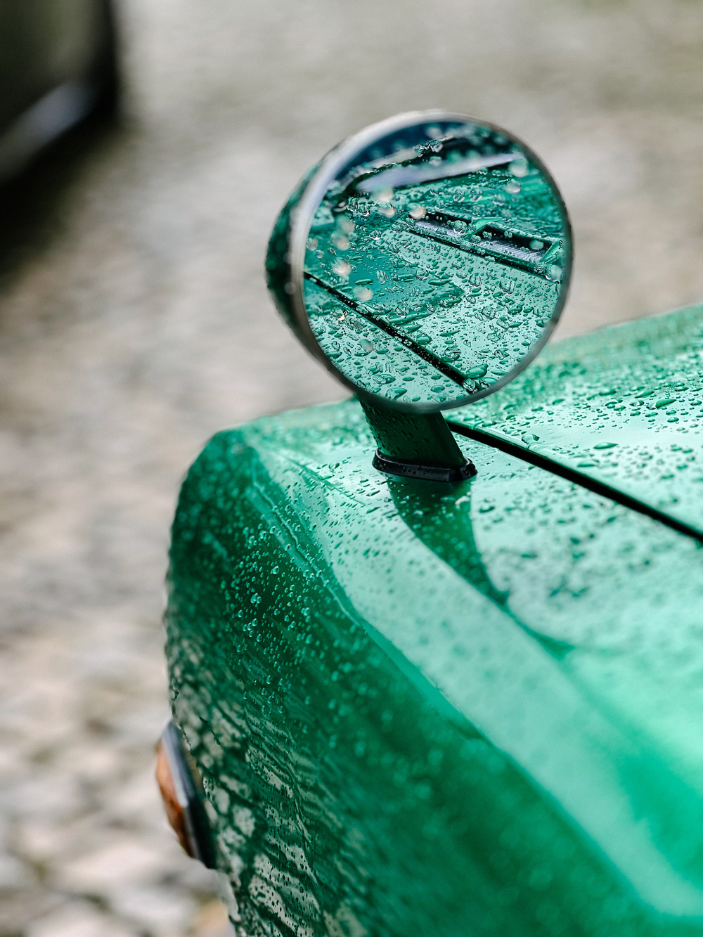 Side mirror and part of a green car covered in raindrops, with a blurred background.