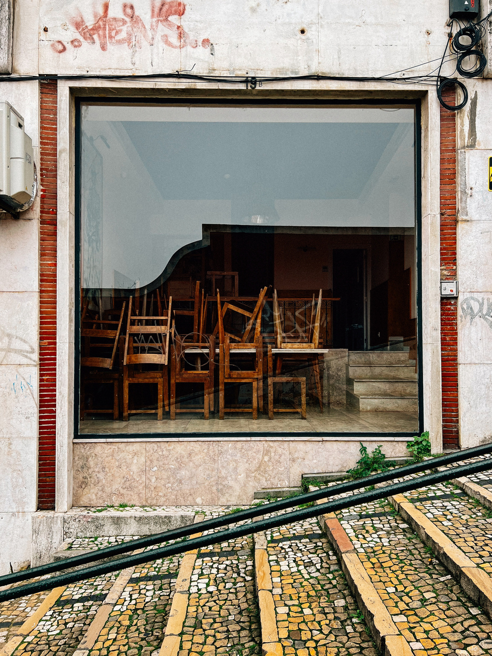 Storefront with large window revealing stacked chairs inside, situated above a sloped cobblestone pavement, and bordered by colorful mosaic patterns.