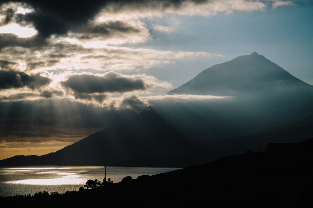 A silhouette of a mountain against a dramatic sky with sun rays piercing through clouds above a body of water.