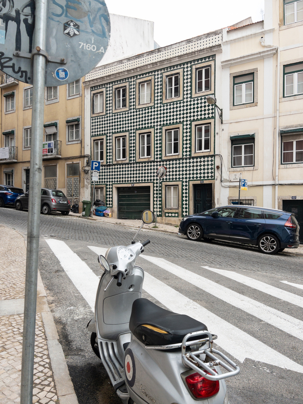 A street corner with a patterned building facade, parked cars, and a silver scooter facing a crosswalk. The building features a distinctive green and white tiled exterior. A street sign and various other buildings are also visible in the background.