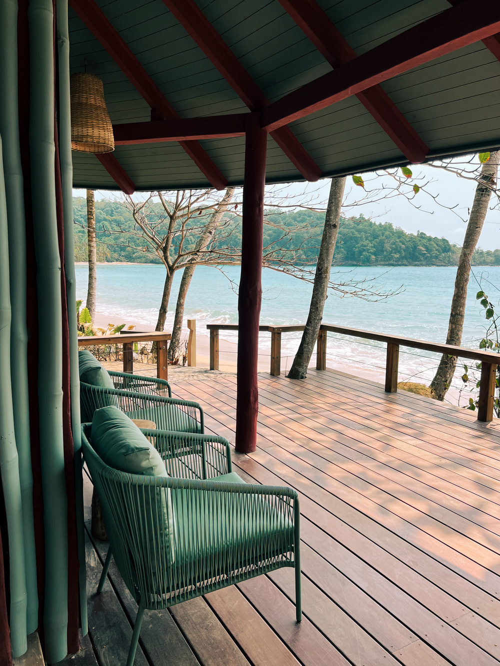 A shaded wooden deck overlooking a serene beach and ocean. The deck features two green cushioned lounge chairs and is supported by wooden beams. Sparse, leafless trees are visible through the railing, and the scene captures a tranquil coastal environment.