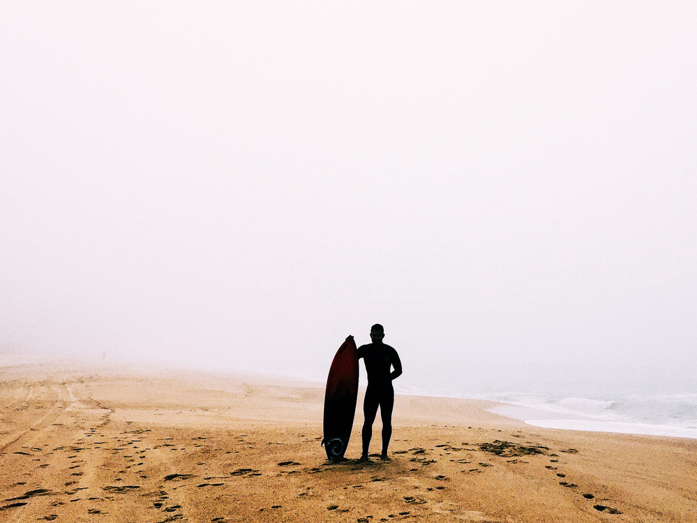 A surfer stands on a misty beach with a surfboard, looking towards the waves.