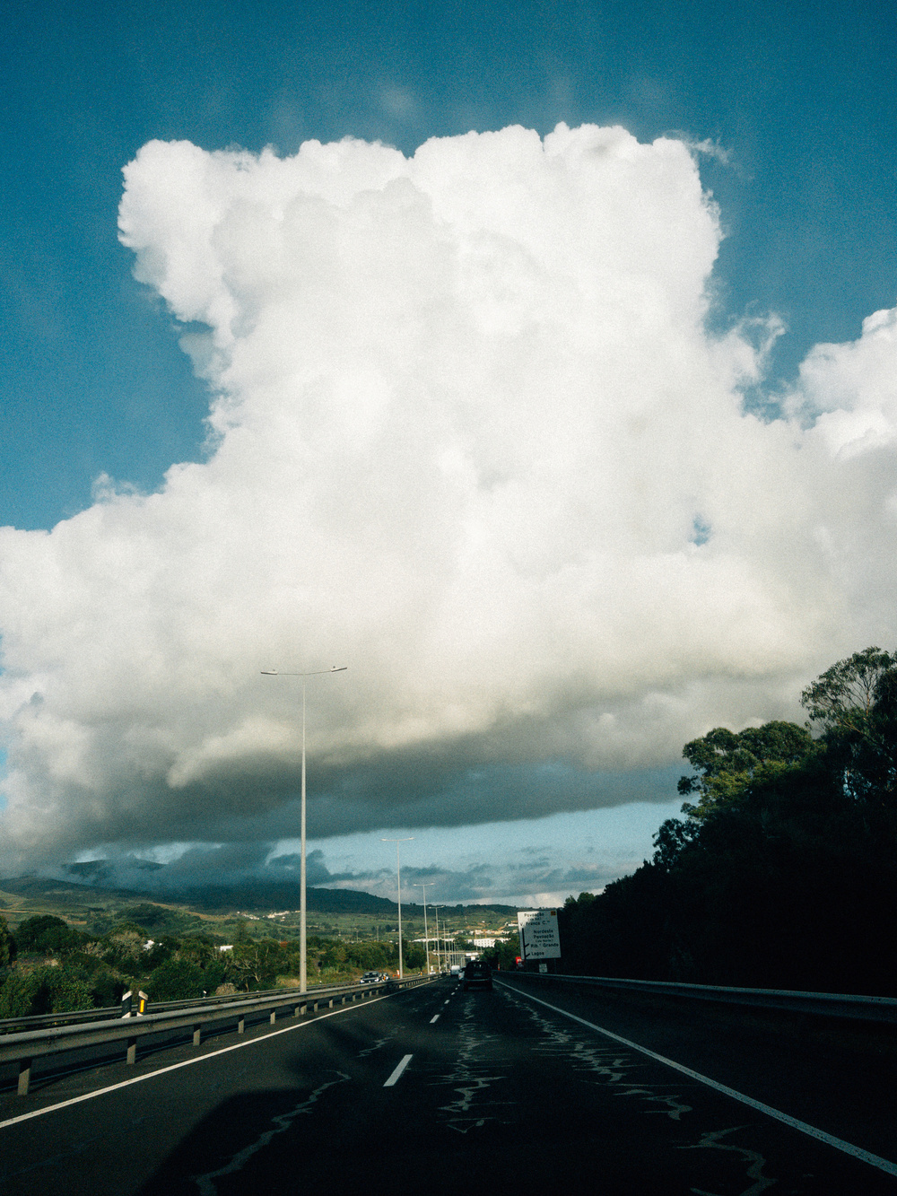 A highway scene featuring vehicles traveling under a massive, white cumulus cloud in the sky. The road is bordered by trees and greenery, and the distant landscape includes hills and scattered buildings.