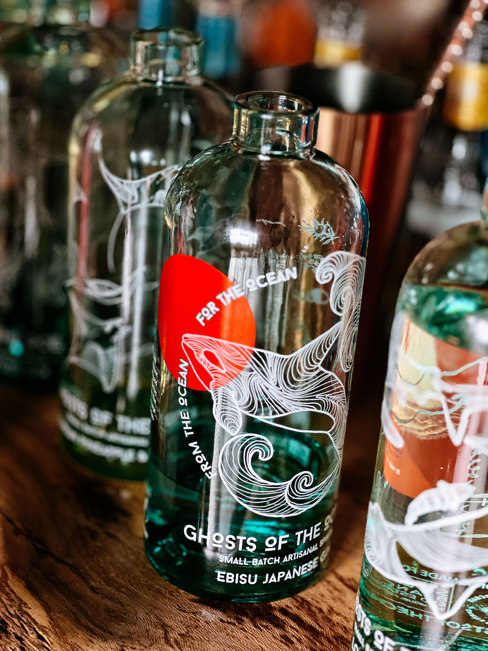 Several glass bottles with intricate wave designs and Japanese text, including the phrases &ldquo;Ghosts of the Ocean,&rdquo; &ldquo;For the Ocean,&rdquo; and &ldquo;Ebisu Japanese.&rdquo; The bottles are placed on a wooden surface and appear to contain a liquid.