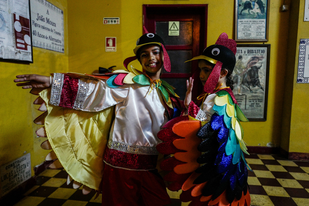 Two children dressed in colorful bird costumes, with sequins and feather-shaped elements, standing in a room with yellow walls and checkered floor tiles, appear to be ready for a performance or celebration.
