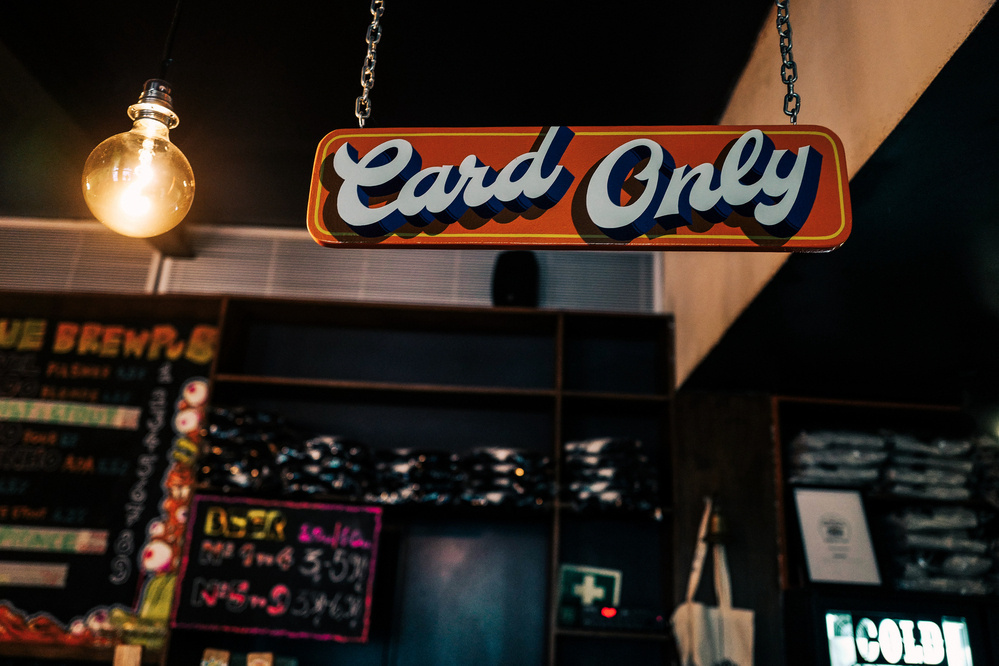 A sign reading &ldquo;Card Only&rdquo; hanging from the ceiling in a cafe or bar environment, with an illuminated light bulb nearby and a faint background featuring a beverage menu chalkboard.