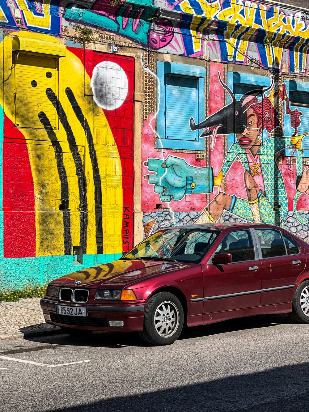 A red BMW car is parked in front of a vibrant, colorful mural on a city street. The mural features abstract designs, including a large yellow shape with black lines, a white circle, and a depiction of a person with red hair.