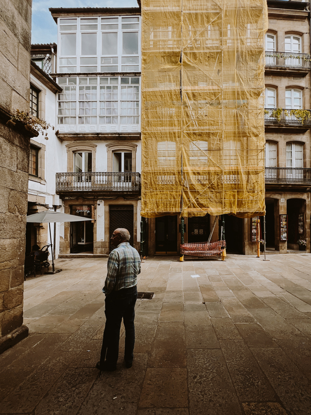 A man stands in a quaint urban square, facing a building partially covered by yellow construction netting. The surrounding architecture features old stone and traditional balconies with ornate iron railings. The area is paved with stone tiles.