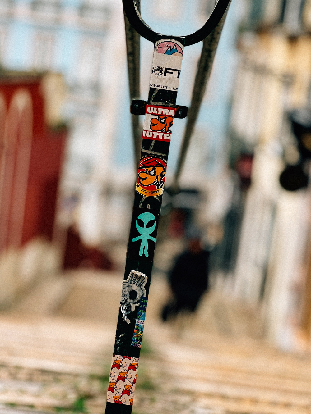 A close-up of a street pole covered in various stickers, with a blurred urban background.