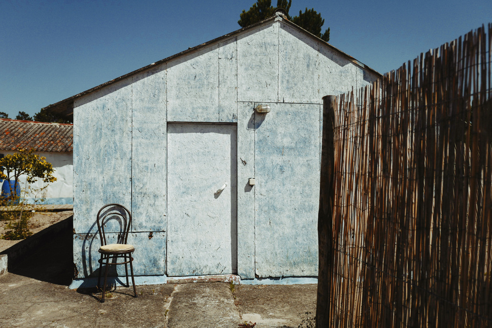 A small, weathered, blue-painted shed with a closed door. An old chair with a cushion is placed against the wall beside the door. A wooden fence and a lemon tree are visible in the background under a clear blue sky.