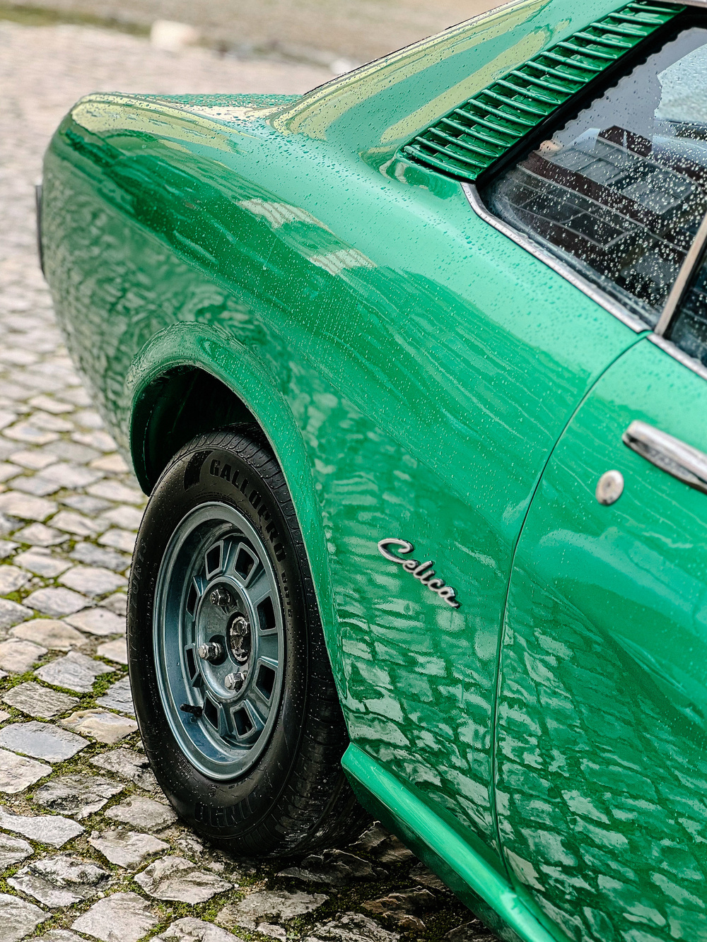 A classic green car with chrome wheels, wet with raindrops, parked on a cobblestone surface. Visible details include the rear side of the car with &ldquo;Celica&rdquo; written on it.
