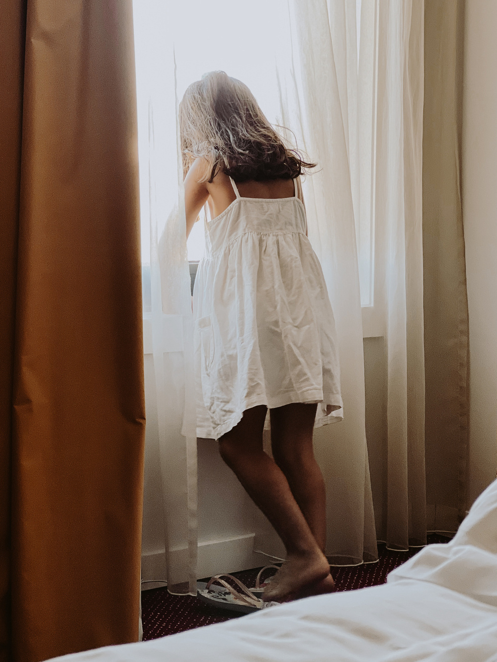 A child wearing a white dress is standing by a window, looking outside. The child is barefoot, and there are light-colored curtains partially drawn around the window. The setting appears to be a bedroom with a bed and yellow curtain visible.