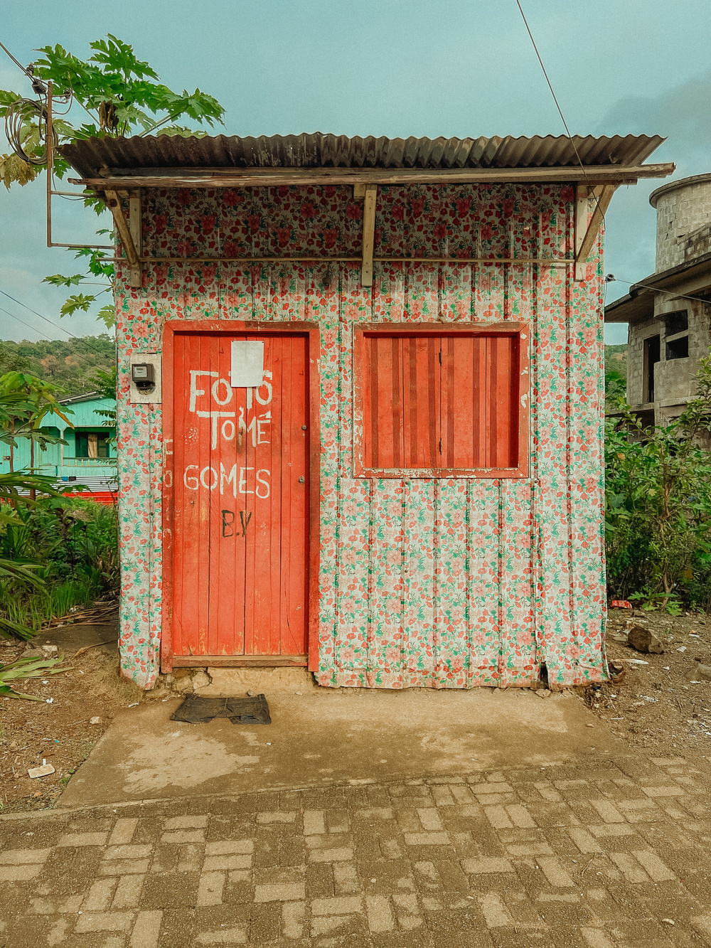 A small, quaint building with floral-patterned exterior walls and red wooden doors and windows. The front door has the words “FOTO TOME GOMES” written in white paint. The building is situated on a paved path with greenery around it. 