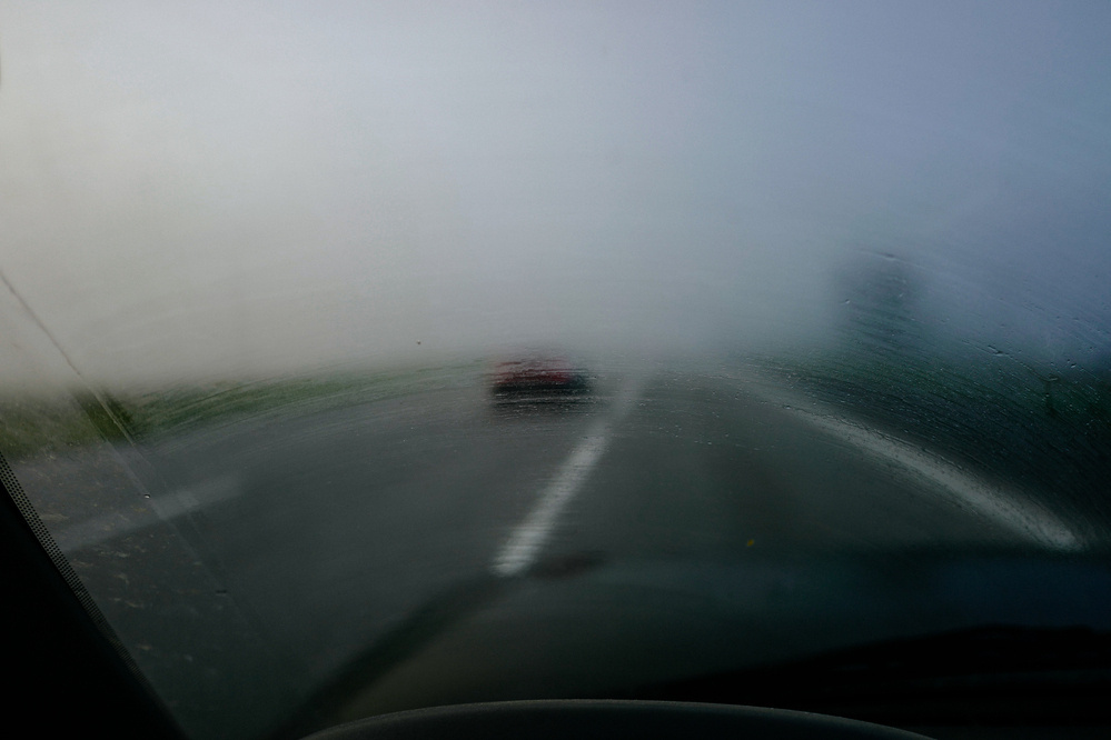 A view through a foggy or misted car windshield showing a blurred vehicle ahead on a road with visible lane markings.
