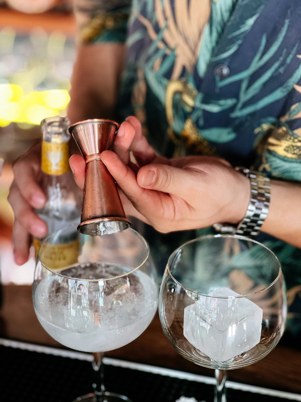 A bartender in a floral shirt and wristwatch is preparing cocktails. He is pouring from a jigger into a glass with ice cubes, and another cocktail glass with a large ice cube is beside it.