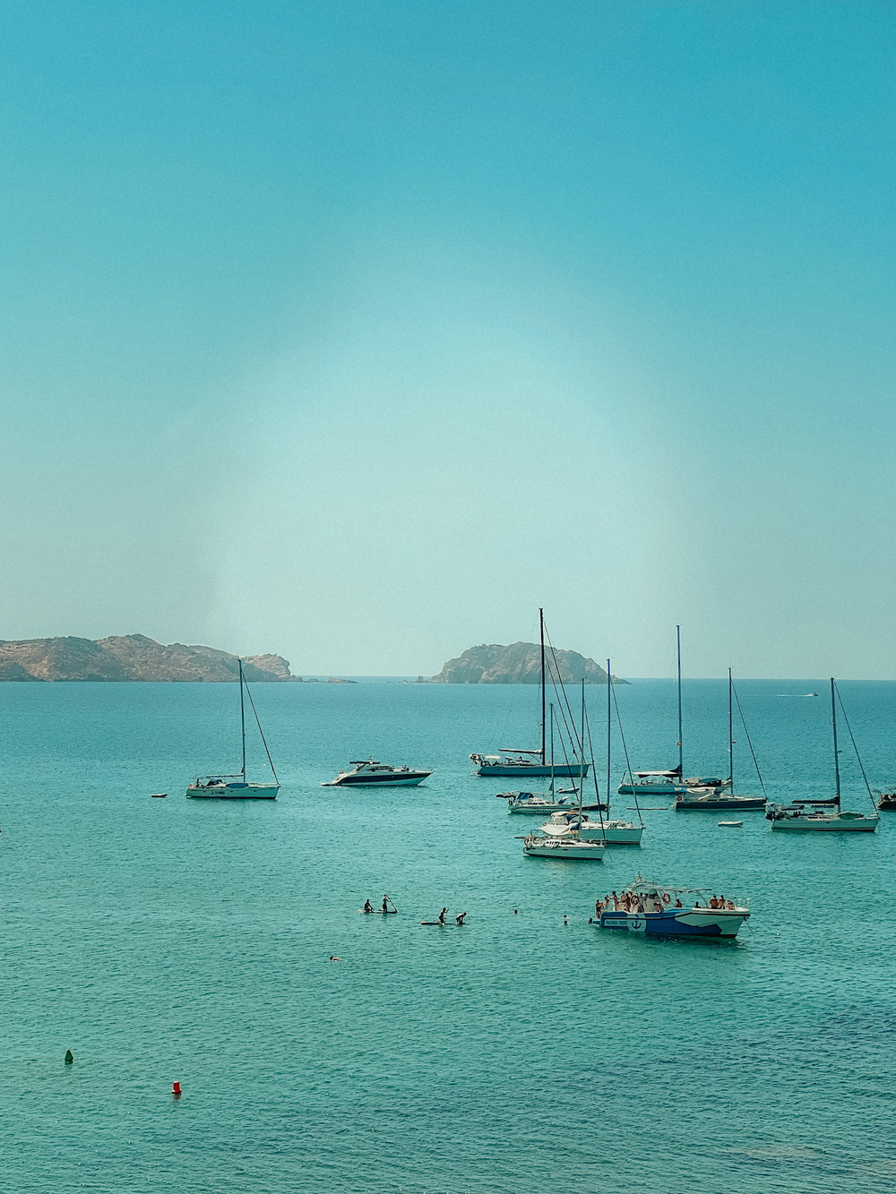 A tranquil coastal scene featuring multiple sailboats and yachts anchored in calm turquoise waters. In the foreground, people are paddleboarding and swimming. In the background, there is a small island and rocky coastline under a clear blue sky.