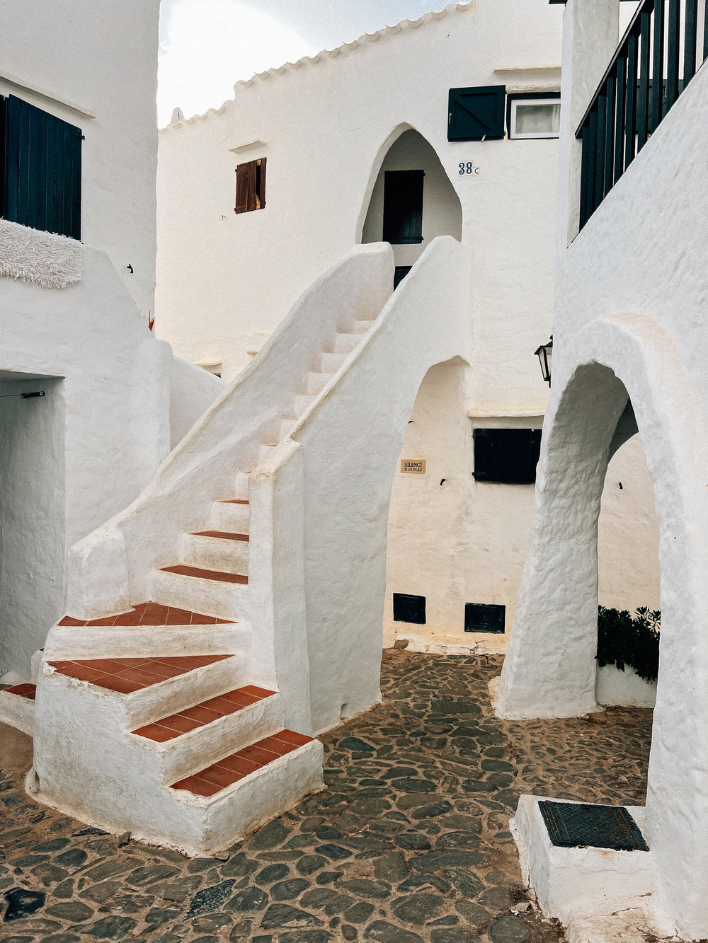 Narrow passageway in a Mediterranean village featuring whitewashed buildings. Prominent elements include a staircase with terracotta steps, arched doorways, and an uneven cobblestone path. Black window shutters and doors contrast against the white walls.