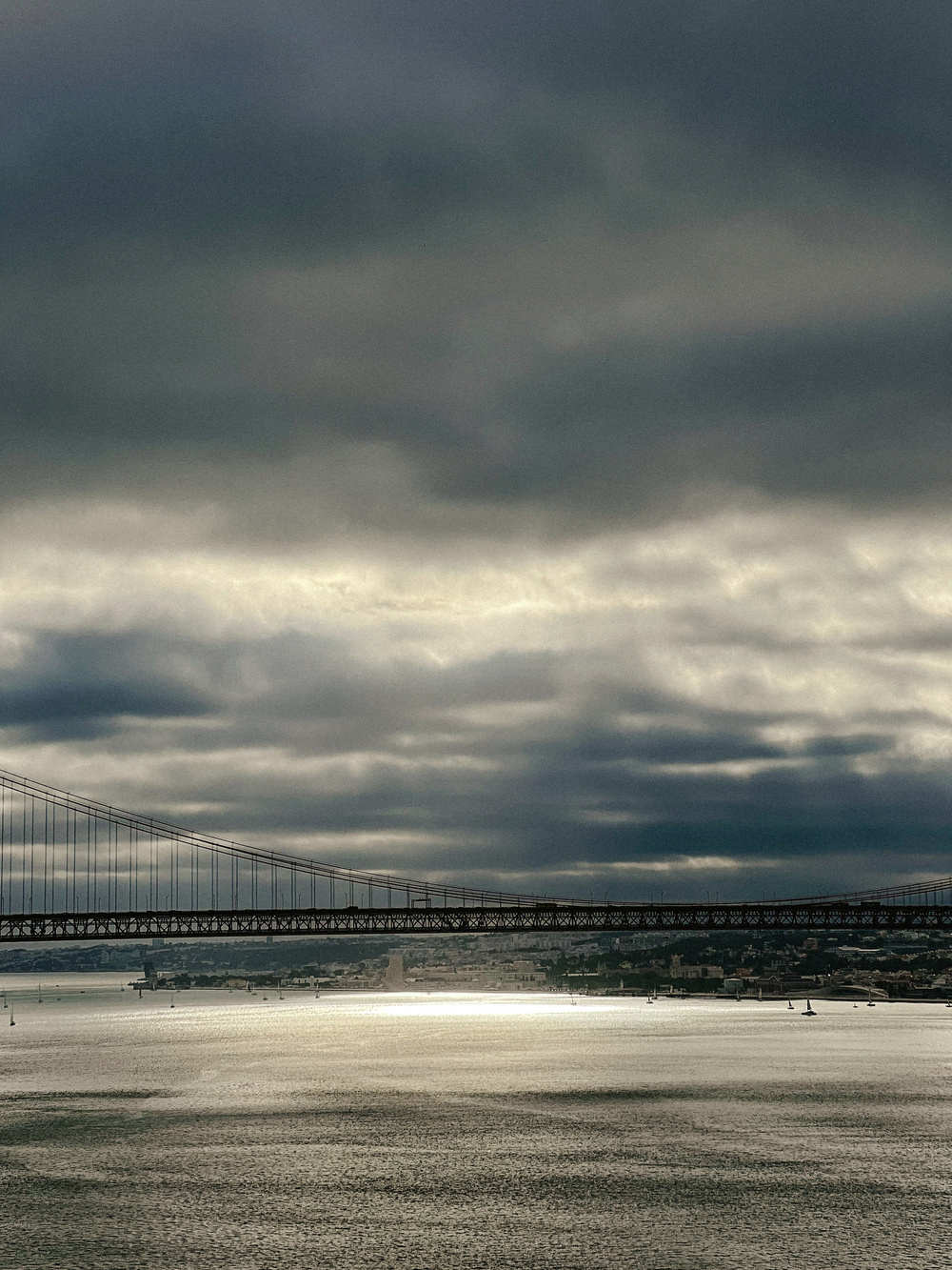 A large suspension bridge spans across a calm body of water under a cloudy, overcast sky. The distant cityscape is partially visible below the bridge, with the sunlight reflecting off the water surface.