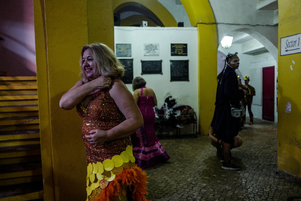 A woman in a sequined dress smiles, standing in an archway. Behind her, various people in costume appear to prepare for a performance or event, in a hallway with yellow walls and plaques.