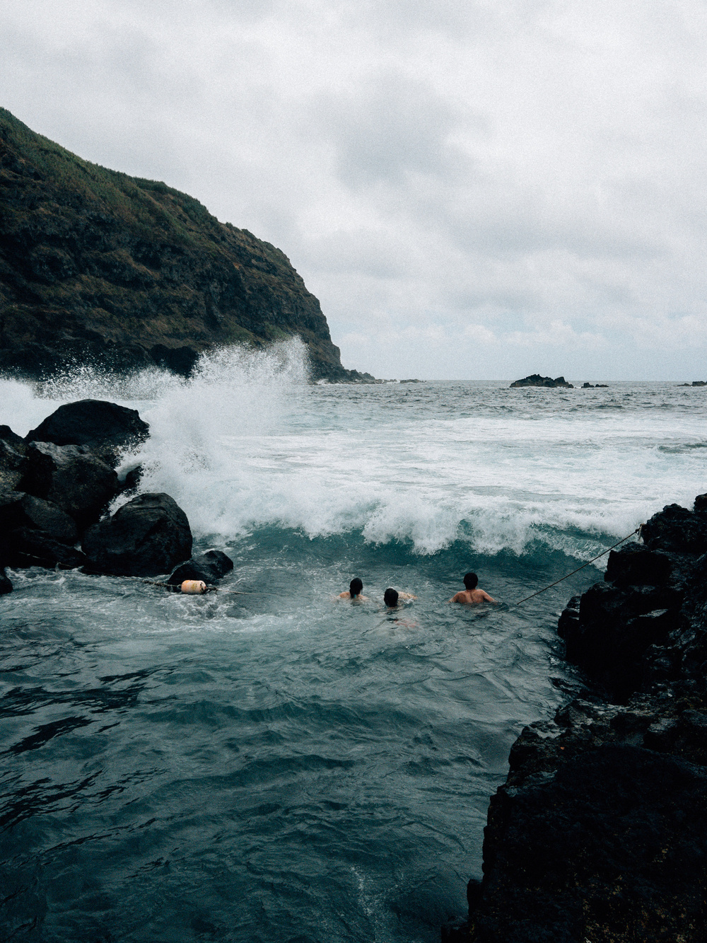 Three people are swimming in rough ocean waters near a rocky coastline. Waves crash against large rocks, and a steep, grassy cliff is seen in the background. The sky is overcast, and the scene has a rugged, adventurous feel.